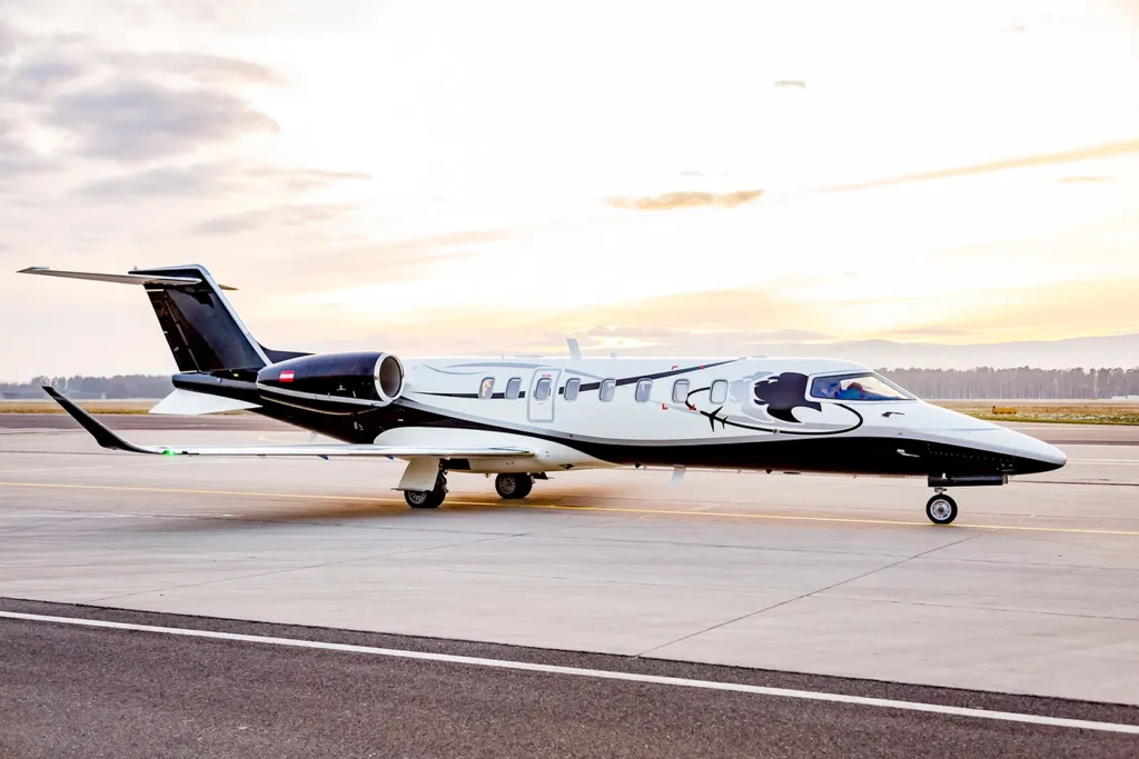 This image features a Bombardier Learjet 75, branded with Avcon Jet livery, parked on the runway during sunset. The jet's sleek design, with its black and white paint scheme and intricate detailing, is prominently displayed from a side angle. The background showcases a soft, glowing sky, adding to the aesthetic appeal of the scene.