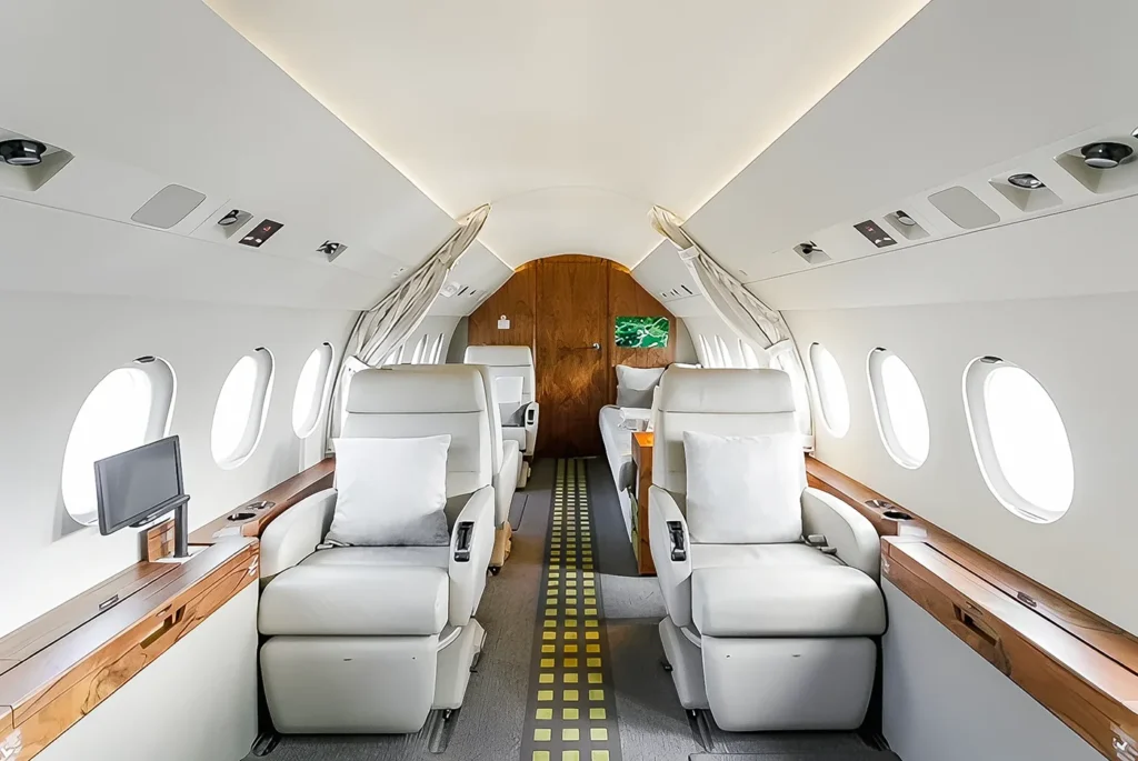 his image shows the interior of a Dassault Falcon 2000LX, showcasing a luxurious and spacious cabin. The soft, cream-colored leather seats are arranged with ample space between them, with wooden accents on the walls and window frames. The cabin is well-lit by multiple large windows, and a small screen is visible near the seats for entertainment. The clean, modern design highlights comfort and elegance, perfect for premium private air travel.