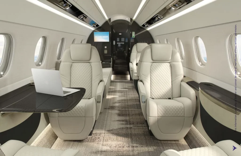 This image shows the luxurious interior cabin of an Embraer Praetor 600, featuring plush, cream-colored quilted leather seats. A fold-out table holds a laptop, highlighting the jet's work-friendly environment. The cabin is well-lit with large windows on the side and sleek overhead storage, offering a spacious and modern ambiance. The image emphasizes comfort and sophistication, ideal for business or leisure travel.