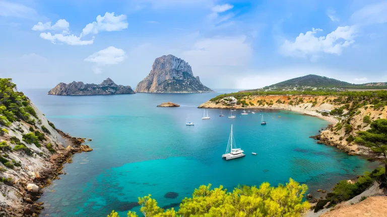Breathtaking panoramic view of the crystal-clear turquoise waters of Ibiza's coastline, showcasing the majestic Es Vedrà rock and a picturesque cove dotted with yachts, serviced by Avcon Jet for exclusive maritime excursions.
