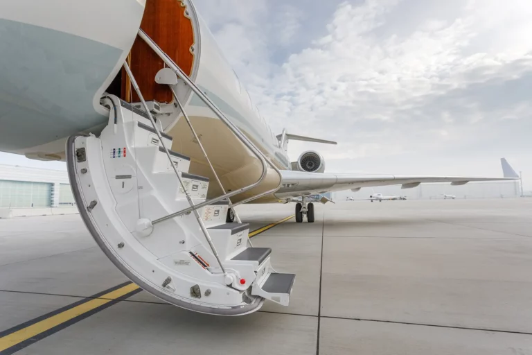 Exterior view of a private jet parked on the tarmac, with its door open showcasing a retractable staircase, suitable for boarding empty leg flights. The aircraft sports a sleek white and silver body under a partly cloudy sky, reflecting a high-end travel experience.